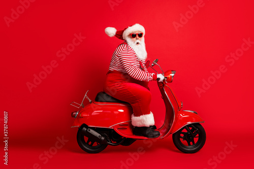 Full length profile photo of excited grandfather grey beard ride retro moped look camera wear santa claus x-mas costume suspenders sunglass striped shirt cap isolated red color background