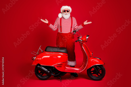 Full length photo of retired grey beard grandpa scooter don't work broken raise hands wear santa x-mas costume suspenders sunglass boots striped shirt cap isolated red color background