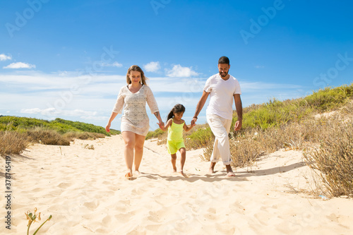 Happy family couple and little kid in summer clothes walking white along sand path, girl holding parents hands. Front view. Family outdoors concept © Mangostar