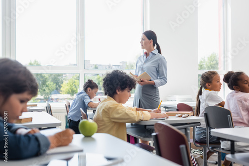 selective focus of teacher holding book near multicultural pupils writing dictation during lesson