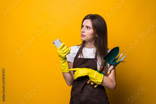 Young womang ardener typing on the phone while holding tools isolated on yellow background