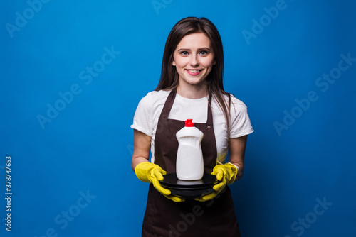 Young woman in apron with washed plates and dish soap against blue background. A bottle of dish soap with a blank label on a stack of clean plates