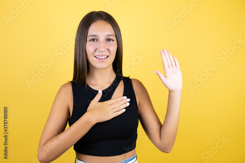Beautiful young girl kid over isolated yellow background smiling swearing with hand on chest and fingers up, making a loyalty promise oath