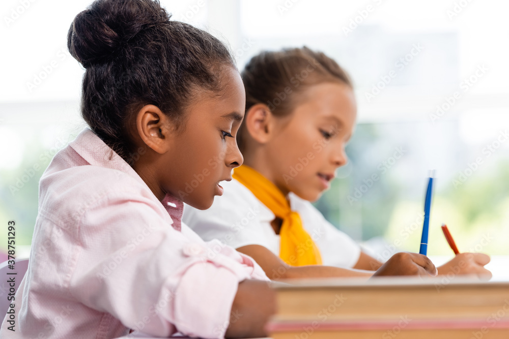 Selective focus of african american schoolgirl holding pen near friend and books in classroom Selective focus of african american schoolgirl holding pen near friend and books in classroom