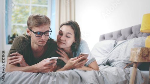 Lovely young happy couple laying on bed using mobile phones and talking