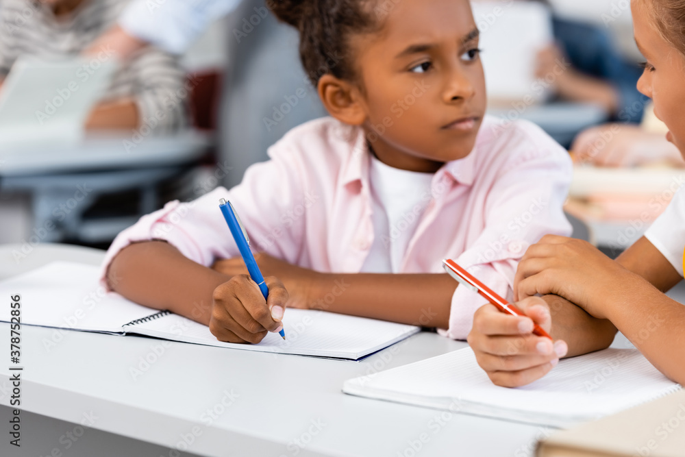 Selective focus of schoolgirl talking to african american friend while writing on notebook in classroom Selective focus of schoolgirl talking to african american friend while writing on notebook in classroom