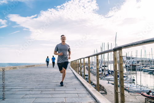 Fototapeta Naklejka Na Ścianę i Meble -  Happy young male runner in tracksuit enjoying active lifestyle on cardio workout near coastline feeling vitality and wellness, positive strong man jogger training flexibility and muscular strength
