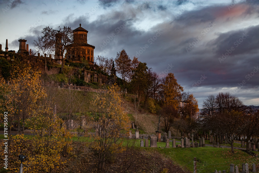 Scotland, Glasgow Necropolis Hill in fall. Grave monuments and mausoleum. Green grass and yellow trees. Yellow light of sunset. Grey cloudy sky. Panorama view. Scotland, Glasgow Necropolis Hill in fall. Grave monuments and mausoleum. Green grass and yellow trees. Yellow light of sunset. Grey cloudy sky. Panorama view.