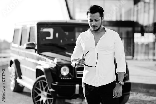 Solid asian man in white shirt and sunglasses posed near black mafia suv car.