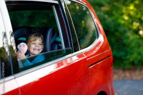 Fotografie Adorable toddler girl sitting in car seat, holding plush soft toy and looking out of the window on nature and traffic