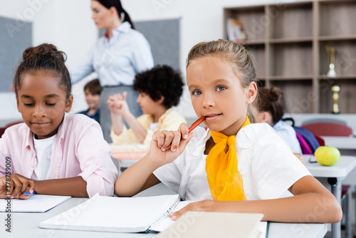 Selective focus of schoolgirl looking at camera near african american friend at desk in school © LIGHTFIELD STUDIOS