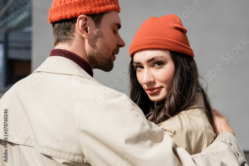 stylish man in trench coat hugging woman in beanie hat outside
