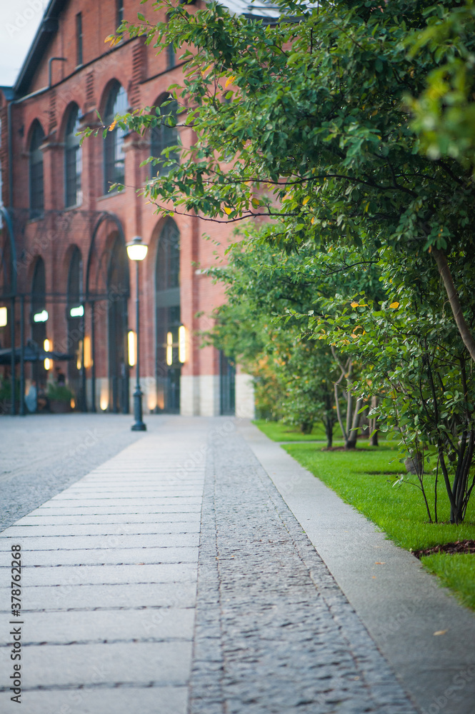 A cobblestone path leading to a large red brick building, a path that runs along green trees. Vertical photo.