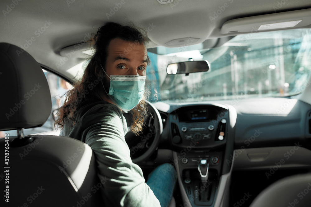 Man taxi driver talking to a passenger while steering the car during coronavirus pandemic wearing sterile medical mask, Social distance and health care concept