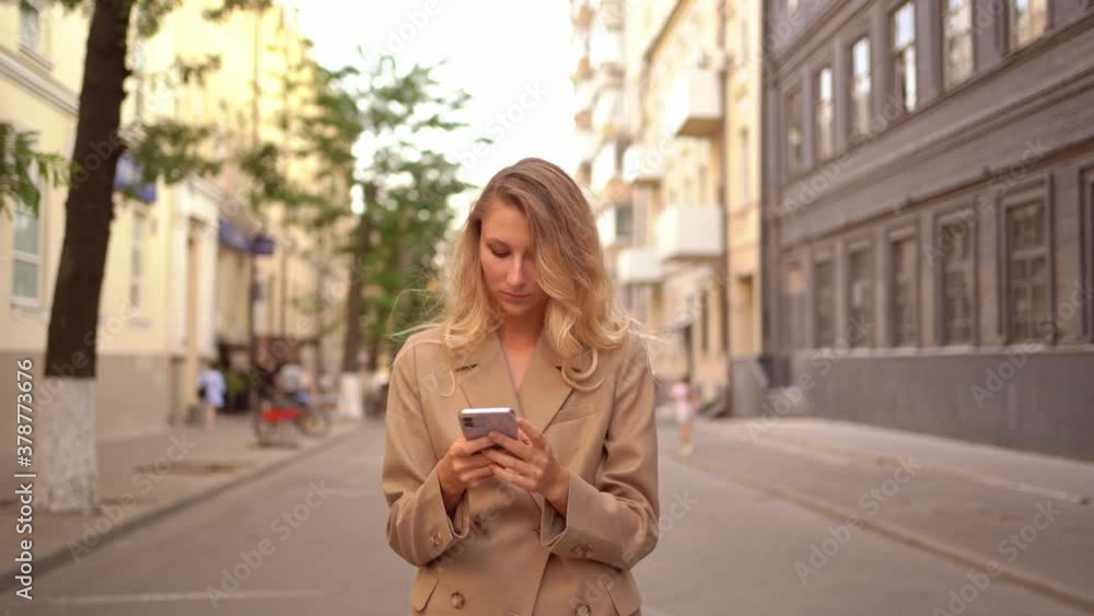 young blonde woman in trench coat with phone in hands is walking down street young blonde woman in trench coat with phone in hands is walking down street