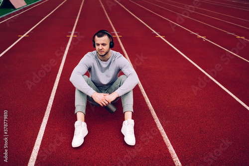 Handsome caucasian male sportsman in active wear resting after training enjoying sound of favorite playlist songs in headphones, pensive hipster guy jogger recreating on stadium listening music