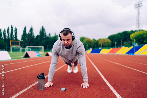 Strong determined caucasian male keeping healthy physical strength on training listening music online via headphones,20s serious sportsman in earphones standing in plank having workout on stadium