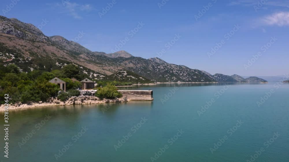 The picturesque shoreline of lake Skadar in Montenegro with tall mountains looming above, trees, stony beaches and cottages around and a stone pier emerging from the coast, aerial scenic view 4K.