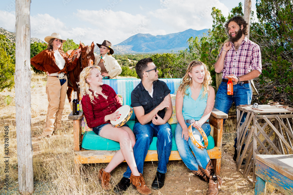 Fototapeta premium group of friends in south western theme in the outdoors camping, drinking, and sitting down on bench enjoying talking, laughing, fun and companionship playing music on a tamborine drum