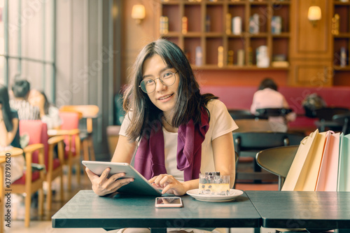 Beautiful young asian woman holding digital tablet and looking at camera with smile while sitting in a modern cafe and near the girl is colorful bags and background look old or vintage style...