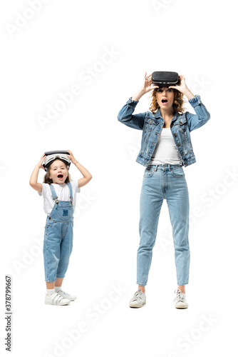 shocked mother and daughter in denim outfits and virtual reality headsets isolated on white