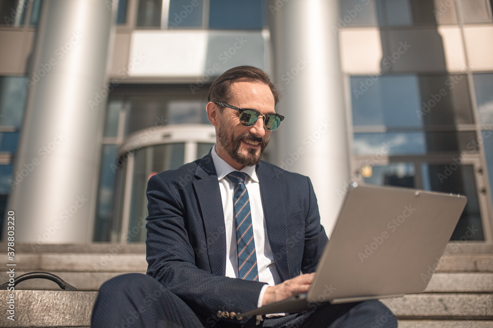Successful businessman works on laptop sitting on steps of business center. Smiling man wearing business suit and sunglasses. High quality photo. Successful businessman works on laptop sitting on steps of business center. Smiling man wearing business suit and sunglasses. High quality photo.