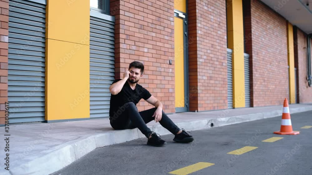 Wide angle with a handsome young Caucasian man talking on the phone while sitting on the curb. Colorful brick background. A young man full of life receives good news over the phone. Wide angle with a handsome young Caucasian man talking on the phone while sitting on the curb. Colorful brick background. A young man full of life receives good news over the phone.