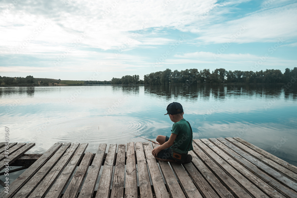 Boy sitting alone on pier while fishing from dock on lake. Kid on beautiful morning in nature