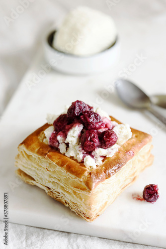 Puff pastry squares with ricotta and cherry sauce on white marble tray. Baked small square cakes topped with cheese and berry sauce
