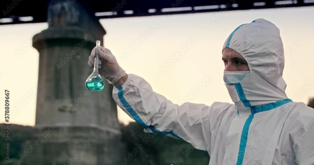 Close up of male chemist ecologist in protective clothing and medical mask taking probes, analysing water samples from dirty polluted river, holding glass tube with chemical agent. Close up of male chemist ecologist in protective clothing and medical mask taking probes, analysing water samples from dirty polluted river, holding glass tube with chemical agent.