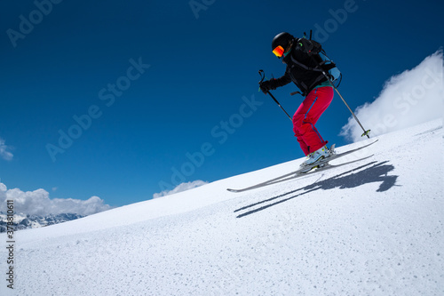 woman skier athlete makes a jump in flight on a snowy slope against the backdrop of a blue sky of mountains and clouds. Freeride and extreme skiing for women