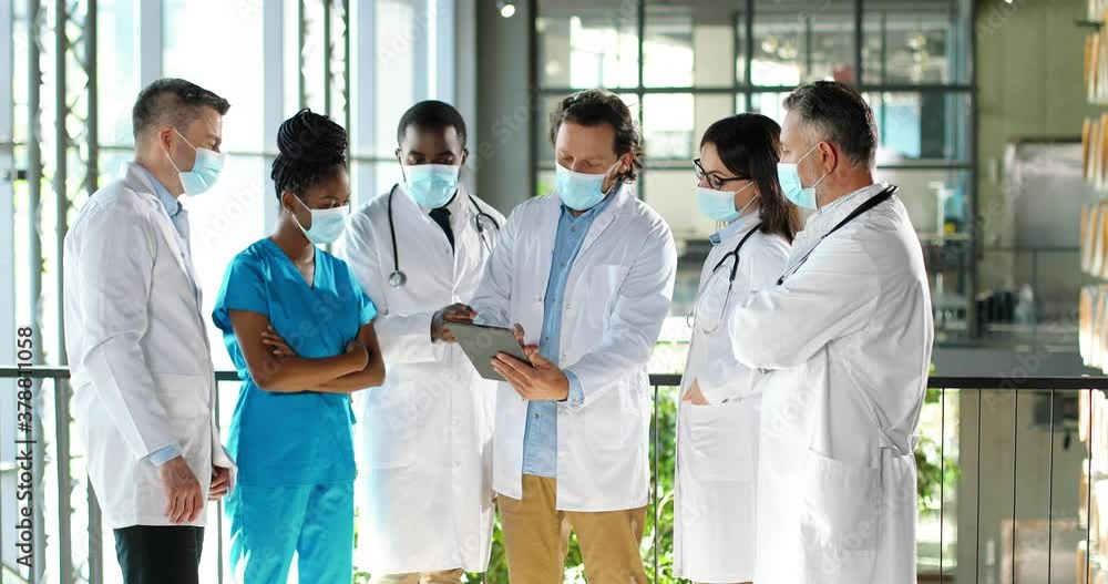Male head of clinic standing with team of mixed-races doctors colleagues and using tablet device while watching on screen. Group of multi ethnic medics, men and women talking and having discussion.
