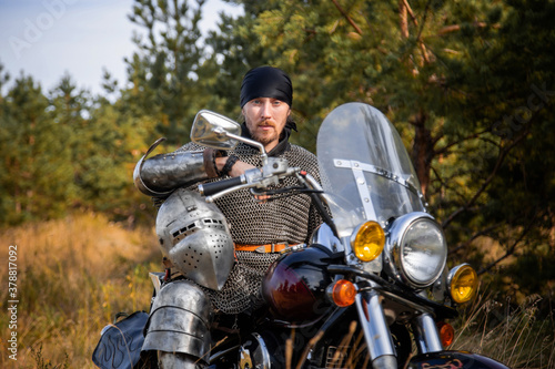 A biker in medieval knight's armor sits on a motorcycle against the background of the forest.