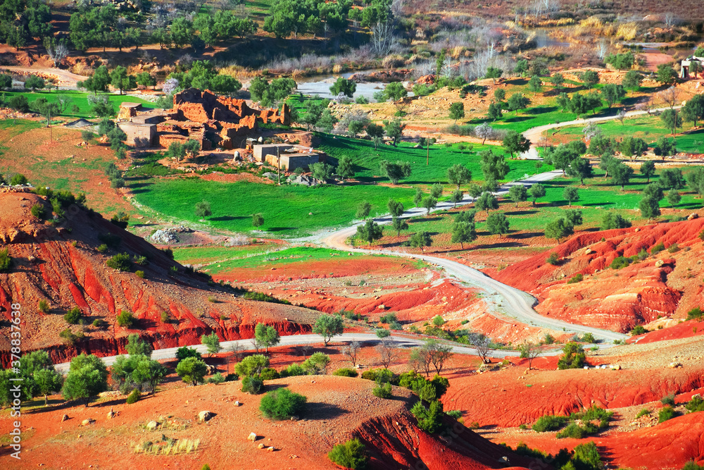 Draa-Tafilalet region, Morocco, North West Africa. Beautiful nature view, a twisty country road among green fields, trees, red hills and ruined ksar - fortified village, Moroccan clay architecture