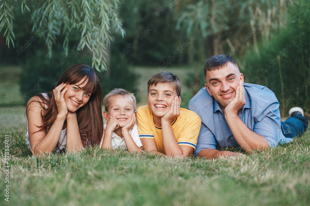 Beautiful happy Caucasian family of four people in park on summer day. Mother and father hugging two sons brothers siblings. Lifestyle authentic natural emotion moment. Beautiful happy Caucasian family of four people in park on summer day. Mother and father hugging two sons brothers siblings. Lifestyle authentic natural emotion moment.