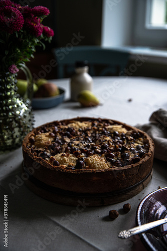 Frangipani tart with pears, hazelnuts and chocolate, plates, fresh pears and vase with flowers on round kitchen table near window.