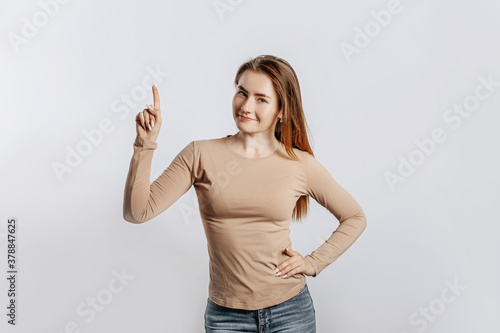 Beautiful young girl smiling and pointing her finger to the side at on a white isolated background. A woman points to an idea, a place for advertising. Positive brunette in a beige jumper.