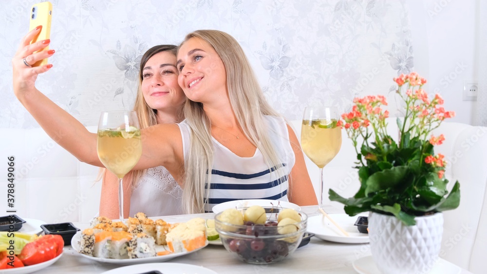 friendship and party concept - two young beautiful girlfriends drink wine and take selfie on smartphone in a cozy kitchen at the table, selective focus friendship and party concept - two young beautiful girlfriends drink wine and take selfie on smartphone in a cozy kitchen at the table, selective focus