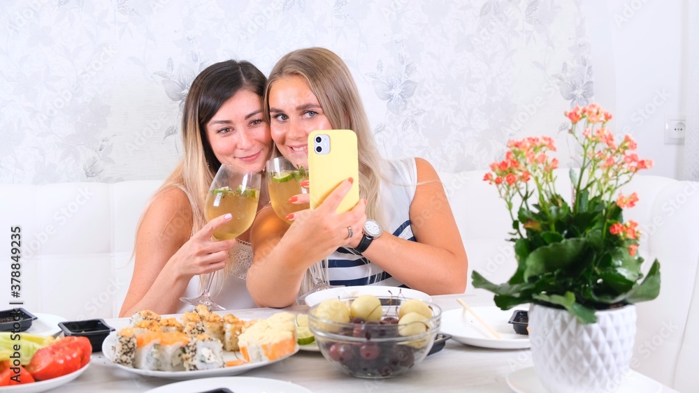  friendship and party concept - two young beautiful girlfriends drink wine and take selfie on smartphone in a cozy kitchen at the table, selective focus