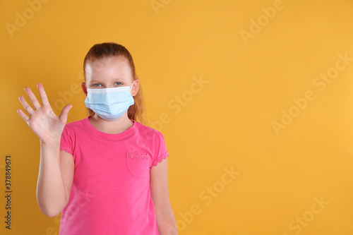 Little girl in protective mask showing hello gesture on yellow background, space for text. Keeping social distance during coronavirus pandemic