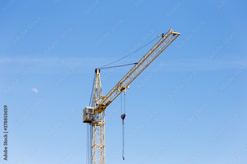 Construction crane tower on blue sky background. Crane and building work in progress. Yellow lifting faucet. Construction concept. Tower crane. Clear blue sky and the moon over the construction crane Construction crane tower on blue sky background. Crane and building work in progress. Yellow lifting faucet. Construction concept. Tower crane. Clear blue sky and the moon over the construction crane