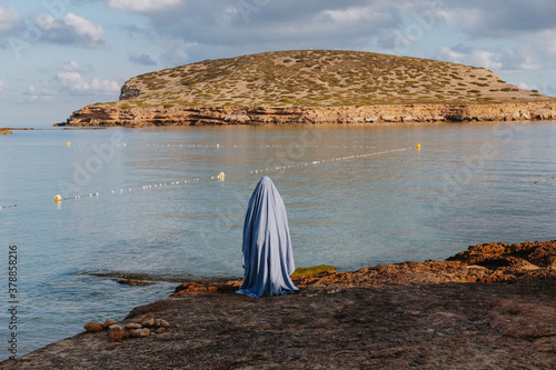Person wearing a ghost costume made from blue sheet  and standing on the Cala Conta beach in Ibiza  photo