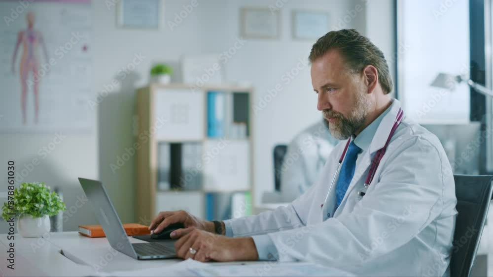 Happy Family Medical Doctor is Working on a Laptop Computer in a Health Clinic. Physician in White Lab Coat is Browsing Medical History Behind a Desk in Hospital Office. 