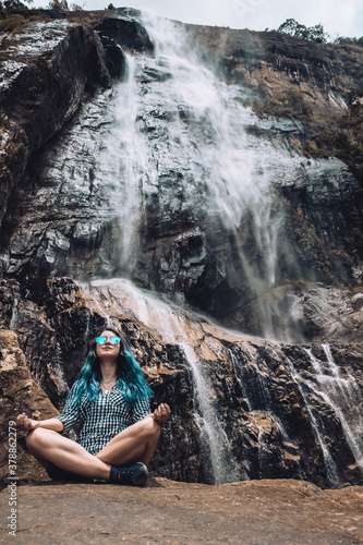 Young white girl sitting under Diyaluma waterfall, Sri Lanka. Happy woman in meditation pose on vacation. Wellness lifestyle, healthy yoga retreat in summer. Peaceful holiday in beautiful exotic place