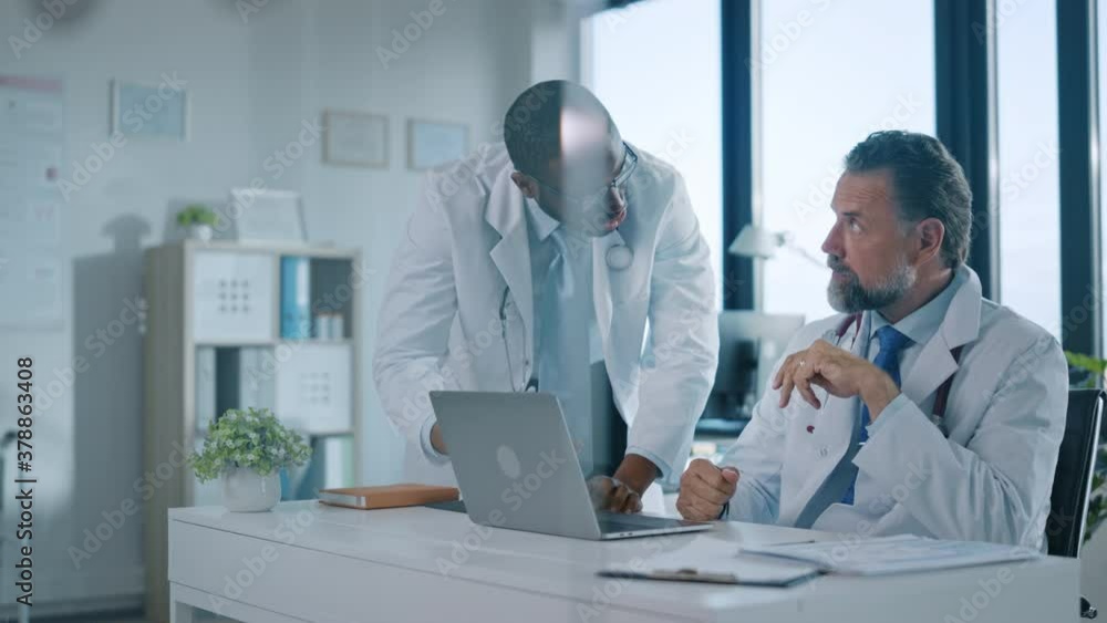 Young African American Physician is Talking to a Senior Doctor in a Medical Health Clinic. Colleagues Discuss Work on Laptop Computer in Hospital Office. Assistant Asks Medical Advice from a Doctor. Young African American Physician is Talking to a Senior Doctor in a Medical Health Clinic. Colleagues Discuss Work on Laptop Computer in Hospital Office. Assistant Asks Medical Advice from a Doctor.