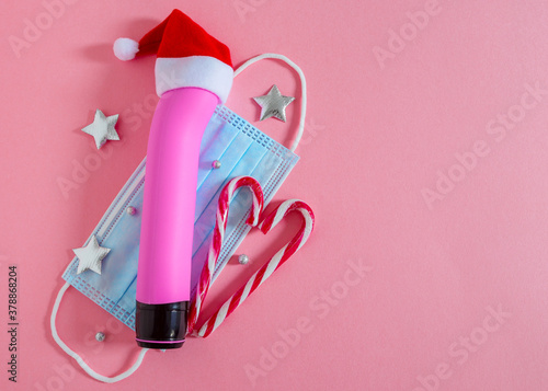 Female dildo (vibrator) with a medical mask and Christmas decor lies on the left on a pink background with a place for text on the right, flat lay close-up. photo