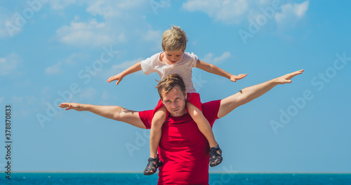 Happy smile father hugs holds on shoulders son boy male child stand blue lazur sea panorama skyline horizon in sunshine day clear sky, play show outstretched hands like airplane. Travel, relationship
