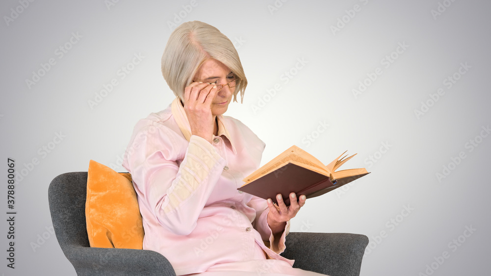Retired woman reading a book sitting on a chair on gradient back Retired woman reading a book sitting on a chair on gradient back