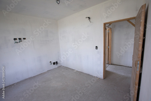 Real estate. House under construction. View of the empty room with white walls, wooden door, concrete floor, and unfinished electrical installations. 