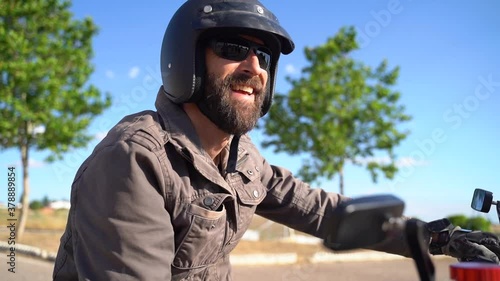 Approaching a dressed beard biker with helmet and sun glasses siting on a motorcycle and smiling. blue sky in background.
